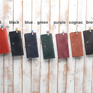 May include: Eight leather wallets hanging on a white wooden wall. The wallets are in the following colors: red, black, blue, green, purple, cognac, and brown.