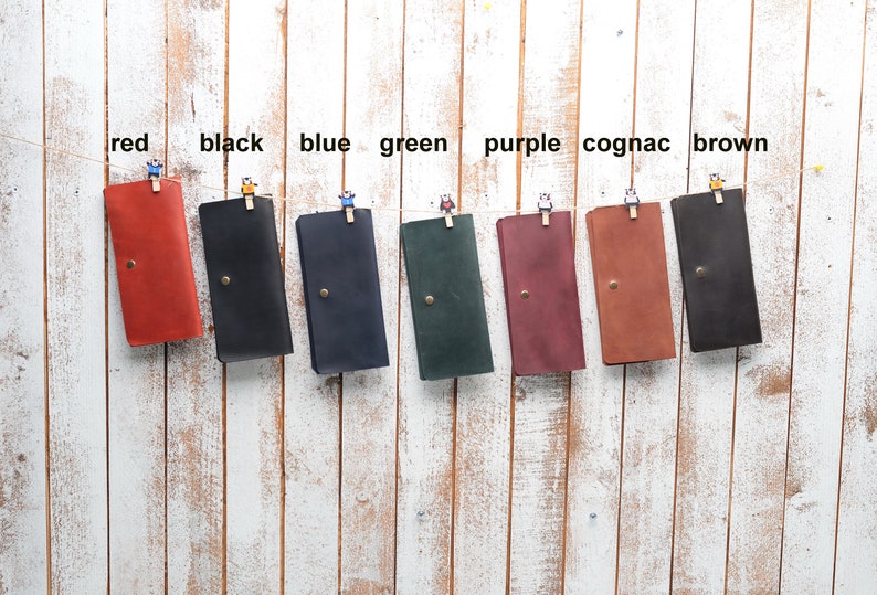 May include: Eight leather wallets hanging on a white wooden wall. The wallets are in the following colors: red, black, blue, green, purple, cognac, and brown.