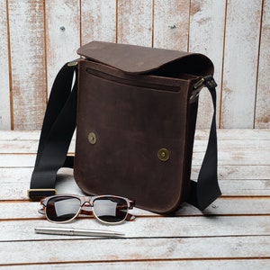 May include: A brown leather messenger bag with a black adjustable strap. The bag is open and has a flap that is folded back. The bag is sitting on a white wooden surface with a pair of sunglasses and a pen.