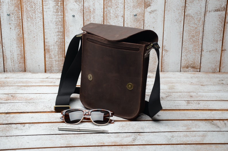 May include: A brown leather messenger bag with a black adjustable strap. The bag is open and has a flap that is folded back. The bag is sitting on a white wooden surface with a pair of sunglasses and a pen.