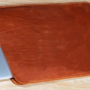 May include: A brown leather laptop sleeve with a stitched edge. The sleeve is open and a laptop is partially inside.