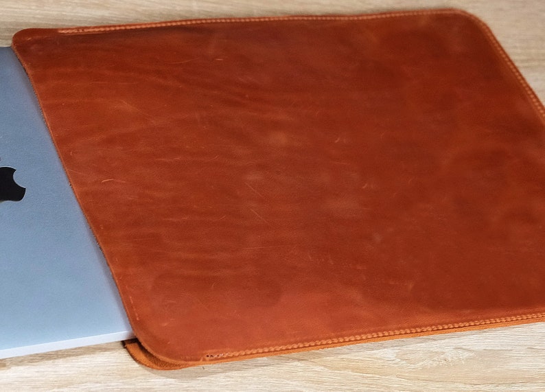 May include: A brown leather laptop sleeve with a stitched edge. The sleeve is open and a laptop is partially inside.