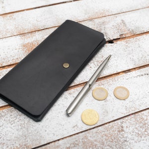 May include: A black leather wallet with a silver pen and three gold coins on a white wooden surface.