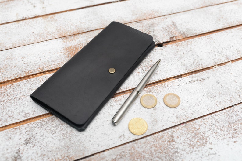 May include: A black leather wallet with a silver pen and three gold coins on a white wooden surface.
