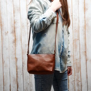 Pu&ograve; includere: Una borsa a tracolla in pelle marrone con una lunga tracolla regolabile. La borsa &egrave; indossata da una persona che indossa una camicia di jeans blu chiaro e jeans.