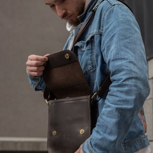 May include: A person wearing a blue denim jacket and a white hoodie is holding a brown leather crossbody bag with a flap and brass hardware. The bag is open and the inside is visible.