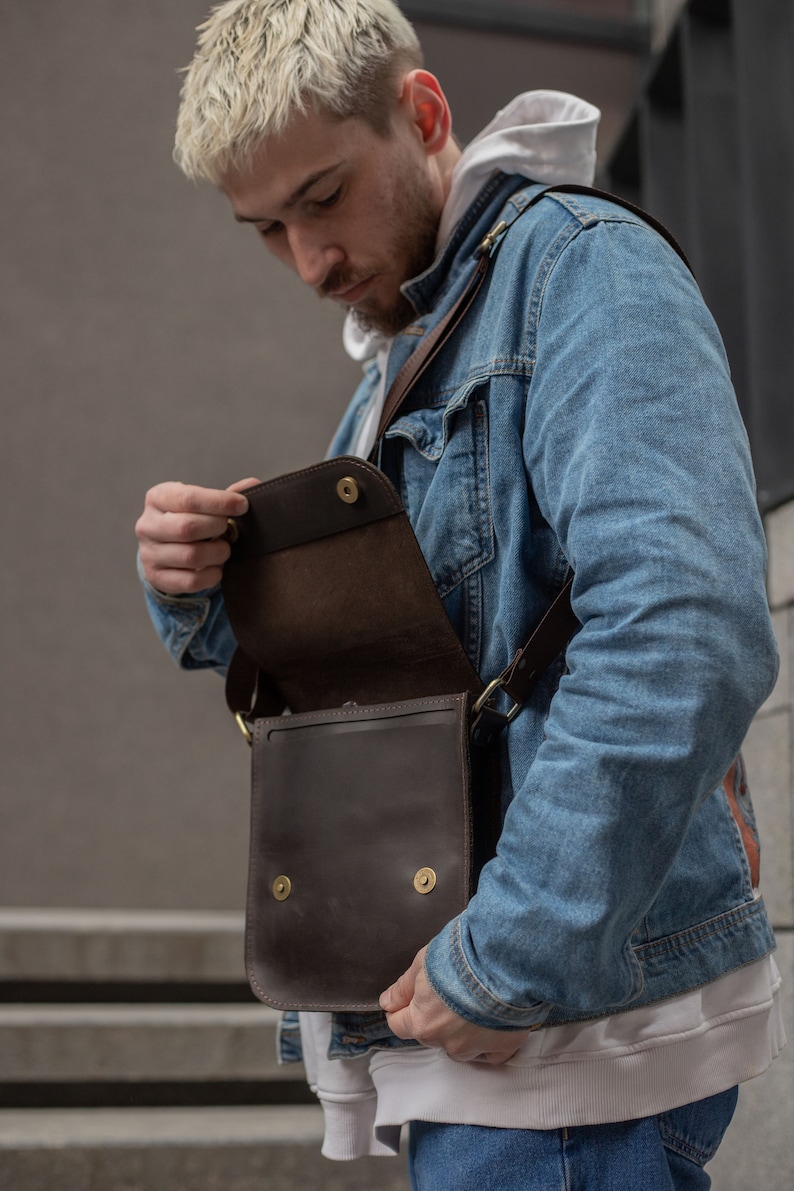 May include: A person wearing a blue denim jacket and a white hoodie is holding a brown leather crossbody bag with a flap and brass hardware. The bag is open and the inside is visible.