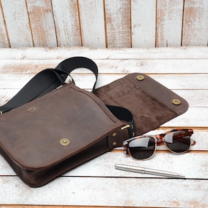 May include: A brown leather messenger bag with a black strap and a flap closure. The bag is open and the inside is lined with brown fabric. A pair of brown sunglasses and a silver pen are lying next to the bag.