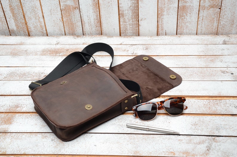 May include: A brown leather messenger bag with a black strap and a flap closure. The bag is open and the inside is lined with brown fabric. A pair of brown sunglasses and a silver pen are lying next to the bag.
