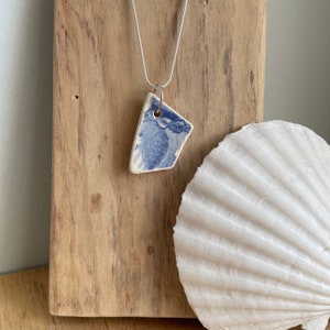 May include: A silver chain necklace with a blue and white ceramic pendant. The pendant is a triangular piece of pottery with a blue and white pattern. The necklace is displayed on a wooden surface with a white seashell.