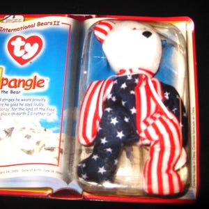 May include: A red, white, and blue Ty Beanie Baby named "Spangle the Bear" is packaged in a plastic case. The bear is wearing a red, white, and blue striped outfit with stars. The packaging has a red, white, and blue Ty logo and the words "International Bears II".
