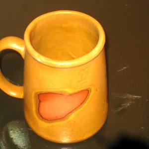 May include: A handmade ceramic mug with a rounded handle and a unique design. The mug is a warm, golden-yellow color with a reddish-orange shape on the front. The mug is perfect for enjoying your favorite hot beverages.