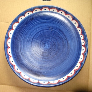 May include: A round, hand-painted ceramic plate with a deep blue center and a decorative scalloped border. The border features alternating white and red accents. The plate's surface has a spiral pattern.
