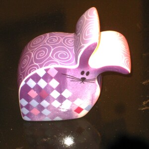 May include: A purple ceramic bunny figurine with a whimsical design. The bunny features a patterned body with a checkerboard design in shades of pink, purple, and white. The ears have a swirl pattern. The bunny has a black nose and whiskers.