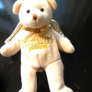 May include: A cream-colored teddy bear plush toy with a gold halo, wings, and bow. The bear has black eyes and a brown nose. The text "Gertrude Hawk Chocolates 2000" is embroidered on the chest in gold.