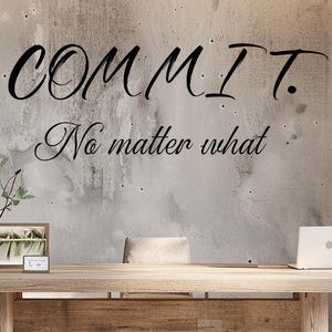 Puede incluir: Oficina moderna con un gran escritorio de madera, un portátil y una estantería. La pared de hormigón presenta el texto "COMMIT. No matter what" en escritura negra. Plantas y lámparas colgantes completan el diseño.