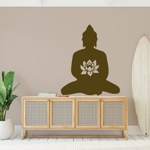 Puede incluir: Silueta marr&oacute;n de un Buda sentado con una flor de loto en una pared beige. Debajo del Buda hay un mueble de madera clara con puertas tejidas. Una tabla de surf blanca se apoya en la pared y una planta en maceta est&aacute; a la izquierda.