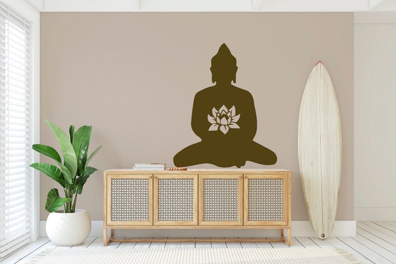 Puede incluir: Silueta marr&oacute;n de un Buda sentado con una flor de loto en una pared beige. Debajo del Buda hay un mueble de madera clara con puertas tejidas. Una tabla de surf blanca se apoya en la pared y una planta en maceta est&aacute; a la izquierda.