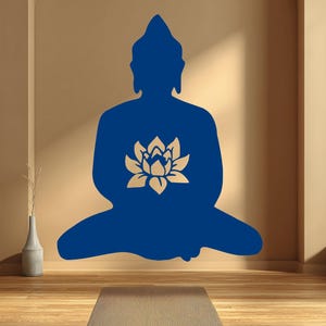 Puede incluir: Silueta azul de un Buda sentado con una flor de loto dorada en el pecho. La imagen est&aacute; en una pared beige con una esterilla de yoga en el suelo de madera. Un jarr&oacute;n con una planta est&aacute; a la izquierda.
