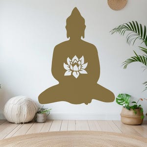 Puede incluir: Una gran silueta dorada de Buda con un dise&ntilde;o de flor de loto blanca est&aacute; fijada a una pared blanca. La imagen es de una habitaci&oacute;n con plantas en cestas y macetas tejidas, un peque&ntilde;o taburete de madera y una alfombra redonda tejida.