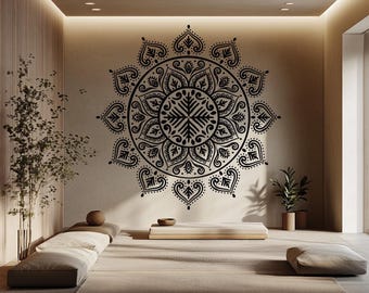 May include: A black mandala wall decal adorns a beige wall in a minimalist room. The intricate design features a central geometric pattern surrounded by ornate, symmetrical shapes. The room includes a low platform, floor cushions, and potted plants, creating a serene atmosphere.