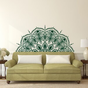 May include: A dark green mandala wall decal above a green sofa with white pillows. The wall art features intricate floral and geometric patterns. Two small wooden side tables with lamps and a vase of flowers flank the sofa.
