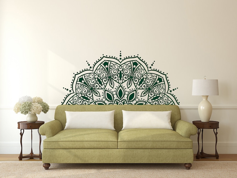 May include: A dark green mandala wall decal above a green sofa with white pillows. The wall art features intricate floral and geometric patterns. Two small wooden side tables with lamps and a vase of flowers flank the sofa.
