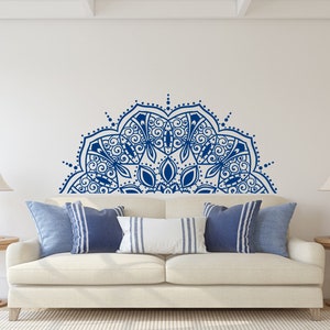 May include: A living room scene featuring a white sofa with blue and white pillows. A large, blue mandala design adorns the wall above the sofa. Two wooden side tables with lamps complete the decor, creating a serene and stylish space.
