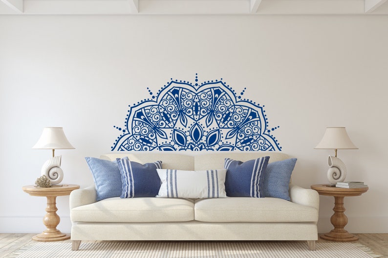 May include: A living room scene featuring a white sofa with blue and white pillows. A large, blue mandala design adorns the wall above the sofa. Two wooden side tables with lamps complete the decor, creating a serene and stylish space.
