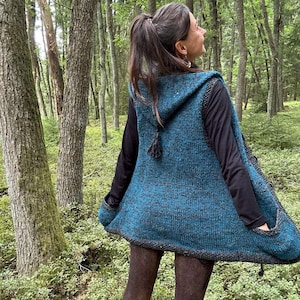 Könnte beinhalten: Eine tealfarbene Kapuzenweste mit einem strukturierten Strickdesign. Die Weste hat eine dunkelgraue Einfassung und Taschen. Die Person trägt ein schwarzes Langarmshirt und braune Leggings. Der Hintergrund ist ein Wald mit hohen Bäumen.