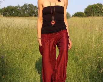 May include: A woman wearing a black tank top and maroon harem pants stands in a field of tall grass. She is wearing a long necklace with a circular pendant.