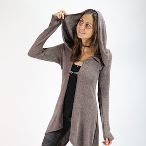 May include: A long, hooded cardigan in a speckled brown fabric. The cardigan has an asymmetrical hemline and a metal clasp closure. The sleeves are long, and the hood is up. The person is wearing black leggings and a black top.