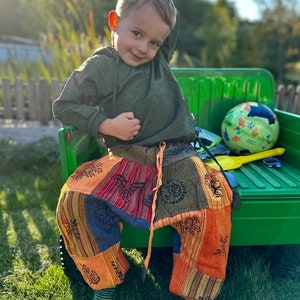 Könnte beinhalten: Ein kleines Kind trägt ein grünes Kapuzenhemd und bunte Patchworkhosen und sitzt auf einem grünen Spielzeugwagen. Das Kind lächelt und schaut in die Kamera. Der Wagen hat eine gelbe Schaufel und einen bunten Ball drin.