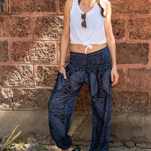 May include: A woman wearing a white tank top with a knotted front and black printed trousers. She is standing in front of a brick wall and looking up.
