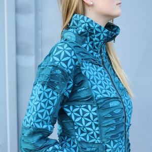 Könnte beinhalten: Türkis- und türkisblaue Jacke mit Reißverschluss und hohem Kragen. Die Jacke hat ein Blumenmuster und dekorative Stoffstreifen. Die Jacke hat einen Frontreißverschluss und Taschen. Die Ärmel sind lang.