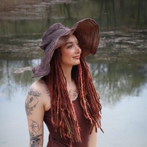 May include: A woman wearing a brown dress and a wide-brimmed hat. Her red dreadlocks are adorned with colorful accents. She is standing near a body of water, with tattoos visible on her arms and legs.