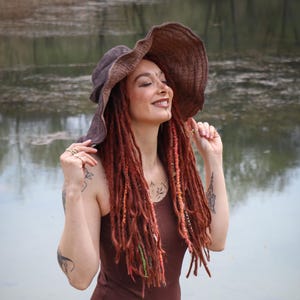 May include: A brown, sleeveless dress with a matching wide-brimmed hat. The woman in the image has long, red dreadlocks and is smiling. The background is a body of water with trees in the distance.