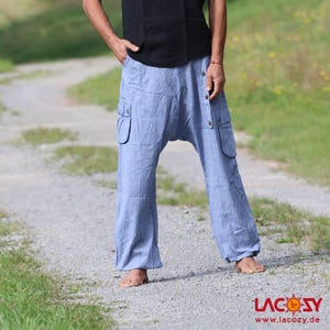 May include: A man wearing a black tank top and light blue loose-fitting pants with cargo pockets. He is standing on a gravel path with green grass and trees in the background.