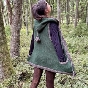 Könnte beinhalten: Eine grüne Kapuzenweste mit grauem Besatz und Quastendetail. Die Weste wird über einem dunkelvioletten Langarmshirt und braunen Leggings getragen. Die Person steht in einem Wald.