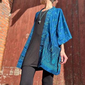 Può includere: Giacca blu stile kimono con motivo paisley e floreale nei toni del verde acqua e turchese. La giacca è indossata sopra un top e leggings neri. Lo sfondo è una porta di legno marrone-rossastro.