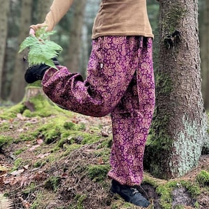 Könnte beinhalten: Eine Person in einem braunen Langarmshirt und einer lila Paisleyhose steht in einem Wald. Die Hose hat ein sich wiederholendes goldenes Muster. Ein lila Schal mit Fransen wird getragen. Die Person hält einen grünen Farn.