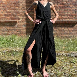 Könnte beinhalten: Ein schwarzer Jumpsuit mit Wickeloberteil und weiten Beinen. Der Jumpsuit hat einen V-Ausschnitt und hohe Schlitze an den Beinen. Das Model steht vor einer Backsteinmauer und grünem Gras.
