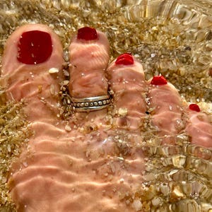 Könnte beinhalten: Nahaufnahme eines Fußes mit rotem Nagellack und einem Silberring, teilweise in klarem Wasser mit Sand und kleinen Muscheln versunken. Die Wasseroberfläche reflektiert das Licht. Das Bild hat eine warme, sommerliche Atmosphäre.