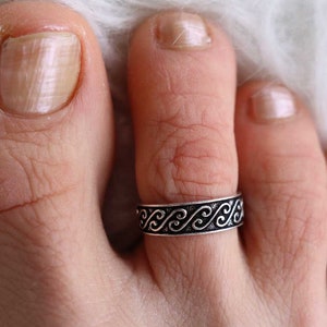 Könnte beinhalten: Ein silberner Zehenring mit einem schwarzen, wirbelnden Design. Der Ring wird an einem Zeh getragen.