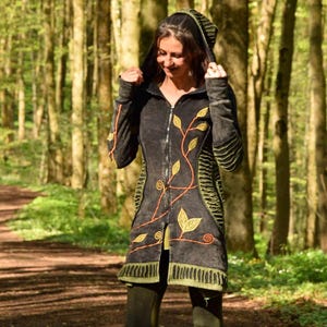 Op de afbeelding: Donkergrijze jas met capuchon en oranje blad- en wijnstokborduurwerk. De jas heeft een ritssluiting en groene accenten. De persoon draagt ​​groene leggings en zwarte laarzen. De achtergrond is een bos met een wandelpad.