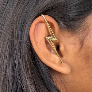 Op de afbeelding: Goudkleurige oor manchet in de vorm van een bliksemschicht, gedragen op het oor. Het sieraad is een uniek accessoire. De oor manchet is een stijlvol en modern sieraad.