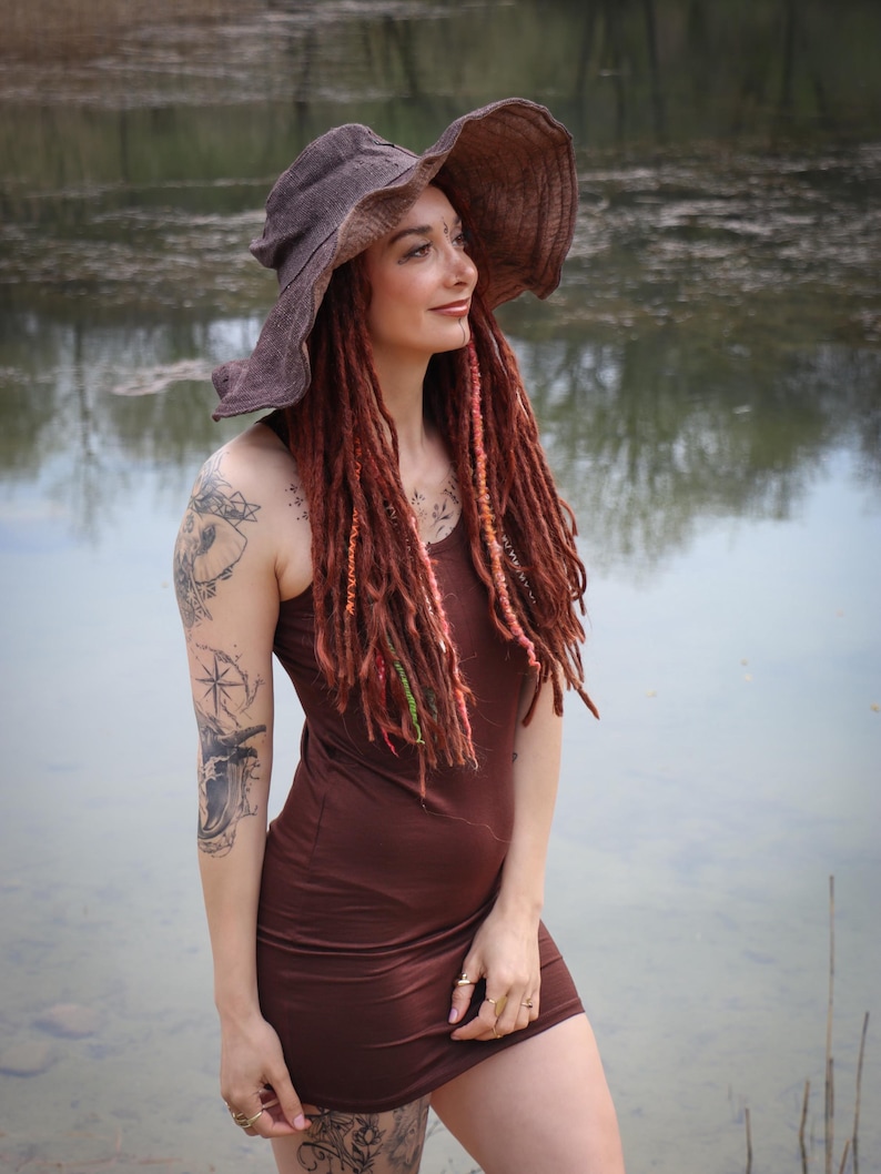 May include: A woman wearing a brown dress and a wide-brimmed hat. Her red dreadlocks are adorned with colorful accents. She is standing near a body of water, with tattoos visible on her arms and legs.