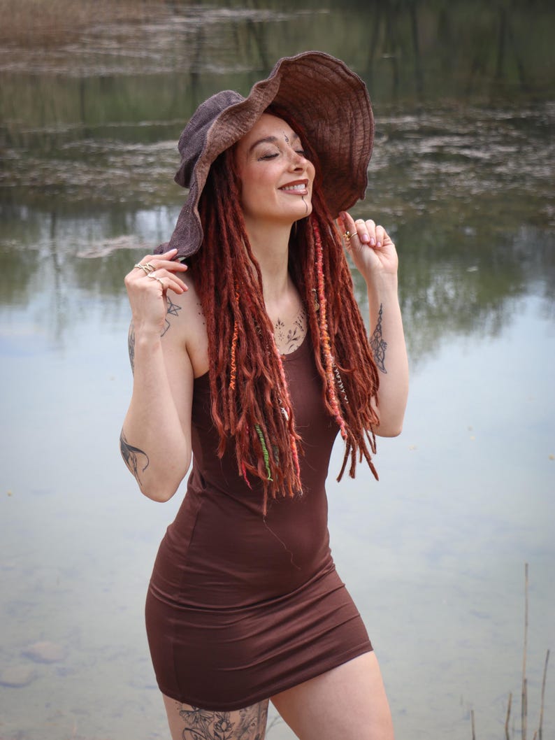 May include: A brown, sleeveless dress with a matching wide-brimmed hat. The woman in the image has long, red dreadlocks and is smiling. The background is a body of water with trees in the distance.