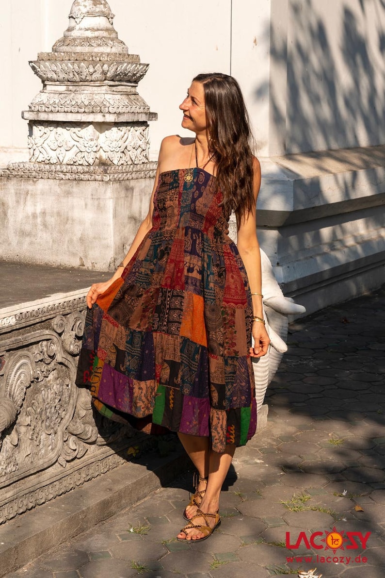 Puede incluir: Una mujer lleva un vestido de retazos marr&oacute;n multicolor con estampado de elefante. El vestido es de tirantes y tiene la cintura fruncida. Est&aacute; de pie frente a una pared blanca con tallas ornamentadas.