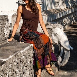 Puede incluir: Una mujer con un top marr&oacute;n sin mangas y una falda de patchwork colorida se sienta en un banco de piedra con una estatua de elefante a su lado. Lleva sandalias marrones con correas de cuero.